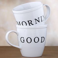 two white coffee cups sitting on top of a wooden table next to each other with the words morning and good written on them