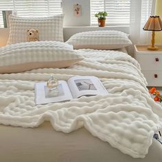 an open book on a bed with white sheets and pillows in front of the window