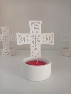 a white candle holder with a cross cut out of it sitting in front of three candles