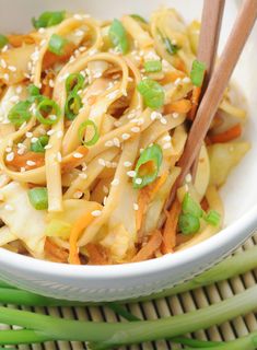 a white bowl filled with noodles and veggies next to chopsticks on a bamboo mat