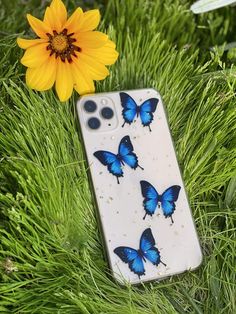 a phone case with blue butterflies on it sitting in the grass next to a yellow flower