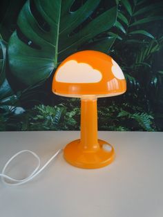 an orange table lamp with a white cloud on it's base and a green leafy background