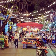 an outdoor market with lots of people and lights strung over the street at night time