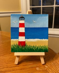 an easel with a painting of a red and white striped lighthouse on it sitting on a wooden table