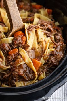 a crock pot filled with pasta and beef, carrots and meat is being stirred by a wooden spoon