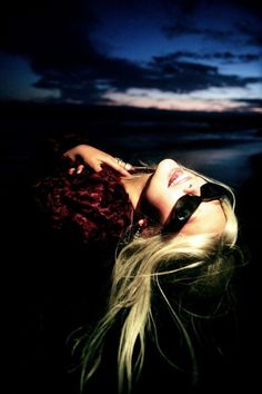 a woman with long hair and sunglasses laying on the ground in front of a body of water