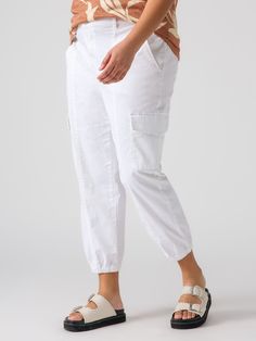 A person wearing a brown-and-beige top is shown from the waist down, dressed in Sanctuary Clothing's Rebel Pant Brilliant White from the Inclusive Collection, featuring side pockets and rolled cuffs, and white sandals with two buckles and black soles. The background is plain white. Jumpsuit Jacket, Utility Pants, High Rise Pants, Military Inspired, Jean Leggings, Outerwear Sweater, Work Pants, Cropped Pants, Outerwear Jackets