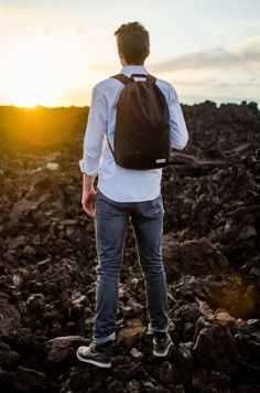 a man with a backpack standing on rocks looking out at the sun setting behind him