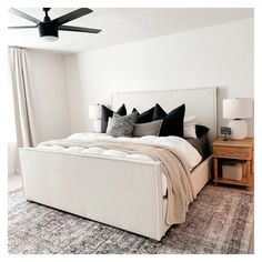 a white bed with black and gray pillows in a bedroom next to a lamp on a dresser