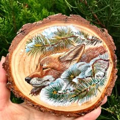 a hand holding a piece of wood with a painting of a fox and pine needles on it