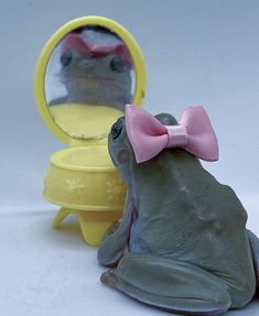 a toy frog with a pink bow sitting in front of a mirror and looking at it's reflection