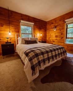 a bed in a room with wooden walls and carpeted flooring next to two windows