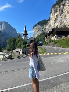 a woman standing on the side of a road in front of some mountains and houses