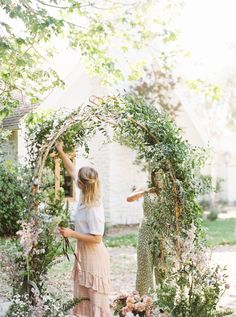 two women standing in front of an arch with flowers and greenery on it,