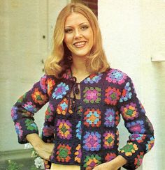 a woman standing in front of a white building wearing a colorful crochet sweater