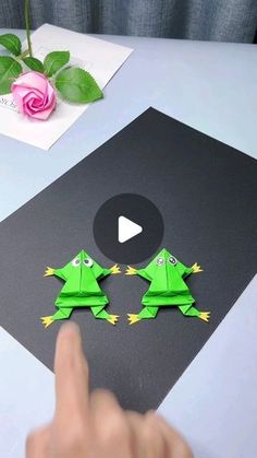 someone is making origami frog decorations on a table with pink rose in the background