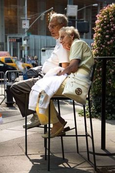 an older couple sitting on top of a metal chair next to each other in front of a building