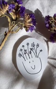 some purple flowers sitting next to a white paper with a drawing on it and a hand