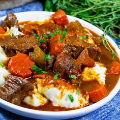 a white bowl filled with meat and vegetables on top of a blue cloth next to some herbs