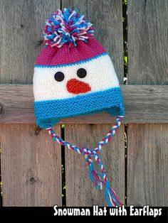 a knitted snowman hat with earflaps hanging on a wooden fence