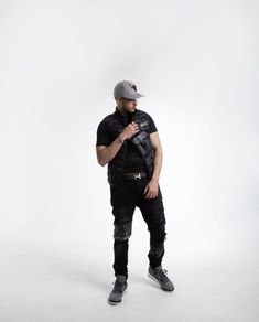 a man standing in front of a white background wearing black pants and a baseball cap