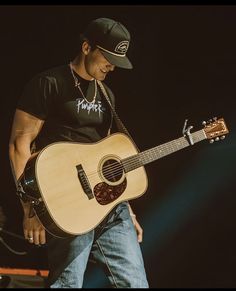 a man holding a guitar on stage at a music festival in the dark with his hat on