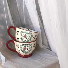 two red and white cups sitting next to each other on a window sill with sheer curtains behind them