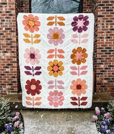 a large quilt hanging on the side of a brick building with flowers in front of it