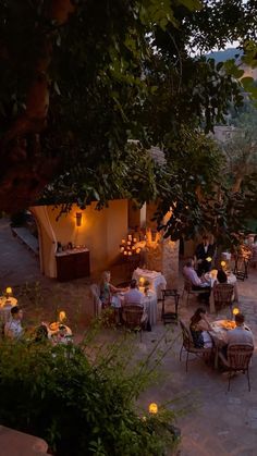 an outdoor dining area with people sitting at tables and candles lit on the tables, surrounded by greenery