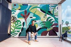 a man sitting on a bench in front of a wall with leaves and plants painted on it