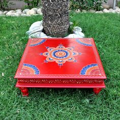 a small red table sitting in the grass next to a tree and stone planter