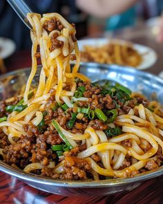 someone is eating noodles with ground beef and green onions in a silver bowl on a wooden table