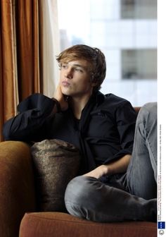 a young man sitting on top of a couch next to a window with his hand under his chin