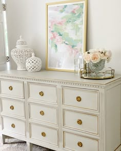 a white dresser with flowers and vases sitting on it's top shelf next to a framed painting