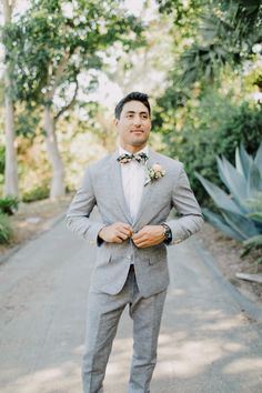 a man in a gray suit and bow tie standing on a road with trees behind him