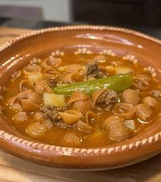 a brown bowl filled with meat and vegetable soup on top of a wooden table next to a green pepper