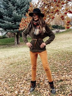 a woman standing in the leaves with her hands on her hips wearing a hat and scarf