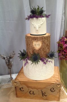 a three tiered cake decorated with purple flowers and greenery sits on a wooden block