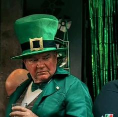 an older man wearing a green top hat and holding a drink in his hand while sitting down