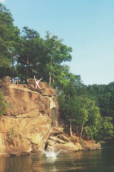 two people jumping off rocks into the water from a cliff near some trees and bushes