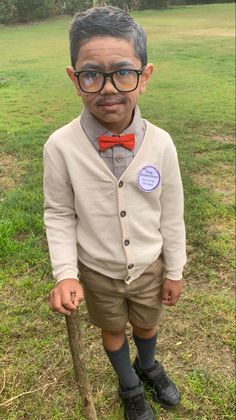 a young boy wearing glasses and a bow tie standing in the grass with a stick