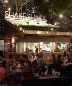 people sitting at tables in front of a restaurant with lights on the roof and outside