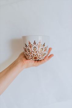 a hand holding a white cup with gold designs on the rim and bottom, in front of a white background