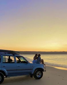 two people are sitting on the roof of a blue car at the beach as the sun sets