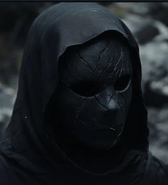 a person wearing a black mask and hood in front of some rocks, with the face of a demon painted on it