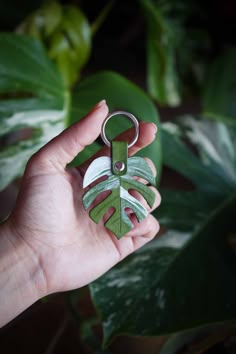 a person holding a green leaf shaped keychain in front of a plant with leaves on it