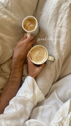 two cups of coffee are being held by someone in bed