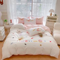 a white bed with pink sheets and pillows in a small room next to a window