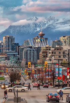 a city street with cars and people crossing the street in front of snow capped mountains