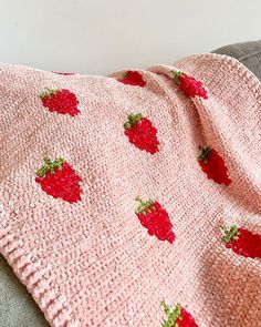 a pink knitted blanket with red strawberries on it sitting on a gray couch